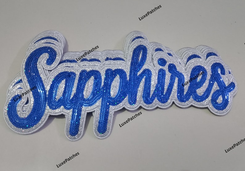 May include: A layered script patch featuring the word Sapphires in bright blue sequins with a white glitter border. The design has a dimensional appearance with multiple stacked layers of white and blue material, creating a thick, raised effect.