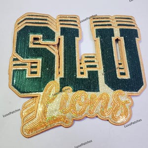 May include: A layered patch featuring the letters SLU in dark green with a glittery gold border, positioned above the word Lions in cursive script. The design uses a multi-layered effect with gold glitter accents and a thick, raised border.