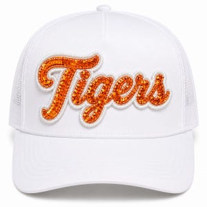 May include: A white trucker hat featuring the word Tigers in orange glittery script lettering across the front panel. The cap has a structured crown, a curved brim, and mesh side and back panels, providing a clean and uniform appearance.