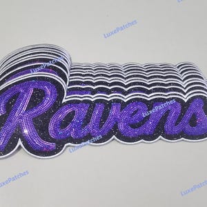 May include: A stack of embroidered patches featuring the word Ravens in a purple script font with a black glitter background and a white border. The patches are layered to create a three-dimensional effect, with the text LuxePatches visible.