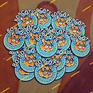 Può includere: Un gruppo di toppe ricamate con un disegno di lince dei cartoni animati. Le toppe presentano le scritte "BOBCATS" e "CHAPMAN HILLS" in un cerchio. I colori principali sono arancione, blu e bianco, con un bordo nero.