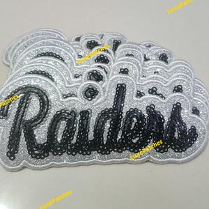 May include: A stack of embroidered patches featuring the word Raiders in black script lettering. Each patch has a silver glitter border and a silver glitter background, creating a layered effect with the black sequin-style text on top.
