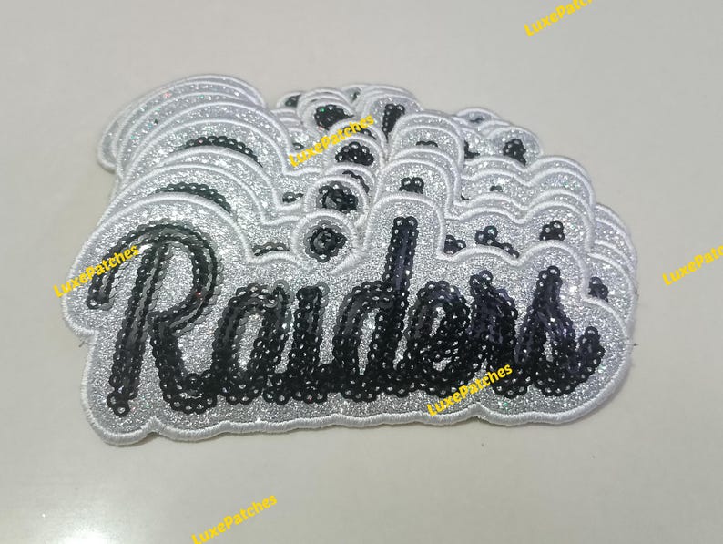 May include: A stack of embroidered patches featuring the word Raiders in black script lettering. Each patch has a silver glitter border and a silver glitter background, creating a layered effect with the black sequin-style text on top.