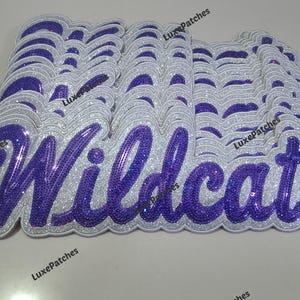 Op de afbeelding: Een stapel geborduurde patches met het woord "Wildcats" in een paars glitterscript. De patches hebben een zilveren glitterrand en een witte achtergrond. De patches zijn waarschijnlijk voor kleding.