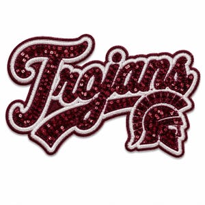 Puede incluir: Un parche decorativo con la palabra Trojans en letra cursiva, adornado con lentejuelas de color granate oscuro y un borde bordado en blanco. El diseño incluye un icono de casco troyano estilizado en el mismo esquema de color granate y blanco.
