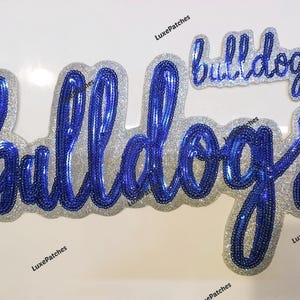 May include: Two sequined patches in the shape of the word "Bulldogs". The larger patch is in a cursive font with blue sequins and a silver glitter border. A smaller patch of the same design is above it.