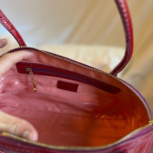 May include: Open view of a red patent leather handbag with a gold zipper and handles. The interior is lined with pink fabric and features a small zippered pocket and a gold charm. The bag is a classic style.