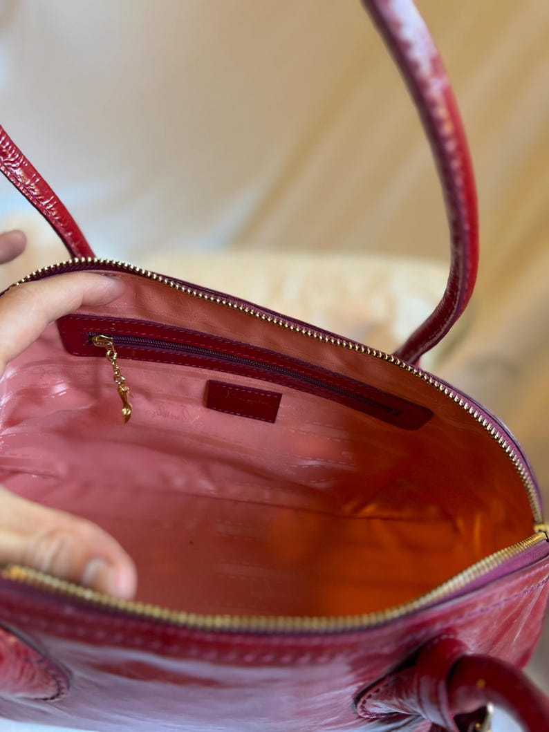 May include: Open view of a red patent leather handbag with a gold zipper and handles. The interior is lined with pink fabric and features a small zippered pocket and a gold charm. The bag is a classic style.