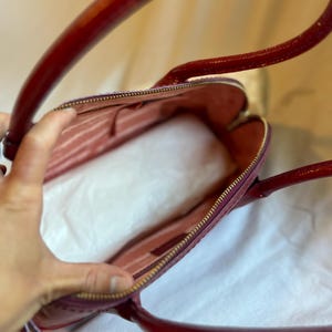 May include: A burgundy handbag with a glossy finish, featuring a gold zipper and matching handles. The interior is a lighter shade of pink, with visible pockets. The bag is open, revealing the inside.