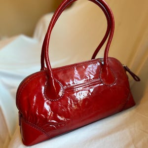 May include: A glossy, red handbag with a rounded shape and two curved handles. The bag features a zipper closure and a small pocket on the back. The patent leather-like material reflects light, giving it a shiny appearance.