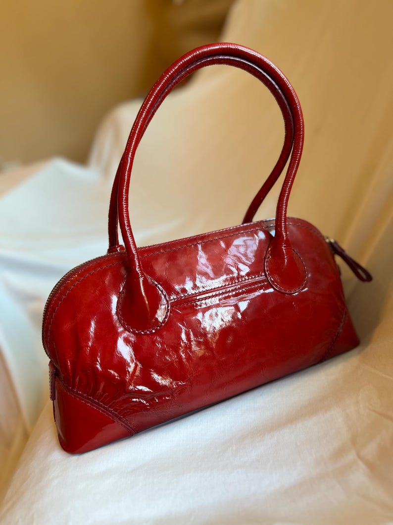 May include: A glossy, red handbag with a rounded shape and two curved handles. The bag features a zipper closure and a small pocket on the back. The patent leather-like material reflects light, giving it a shiny appearance.