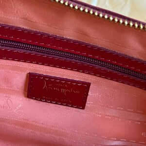 May include: Close-up of a red leather handbag interior. The interior is a light pink color. A small red leather tag with the brand name "Hanamaru" is visible. The zipper and trim are a darker red with gold accents.
