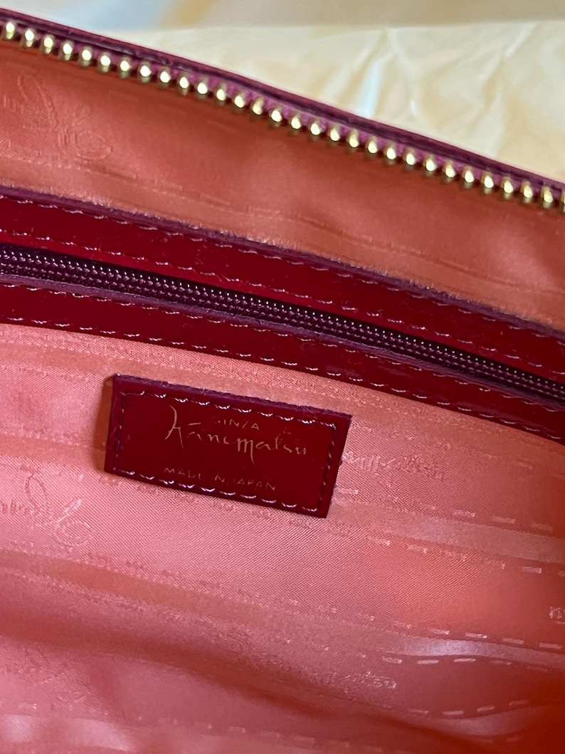 May include: Close-up of a red leather handbag interior. The interior is a light pink color. A small red leather tag with the brand name "Hanamaru" is visible. The zipper and trim are a darker red with gold accents.