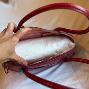 May include: A red, oval-shaped handbag with a glossy finish. The bag has two curved handles and a gold-colored zipper. The interior is lined with a light-colored fabric. The bag is open, revealing the inside.