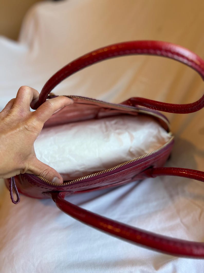 May include: A red, oval-shaped handbag with a glossy finish. The bag has two curved handles and a gold-colored zipper. The interior is lined with a light-colored fabric. The bag is open, revealing the inside.