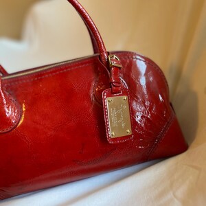 May include: A glossy red handbag with a top handle and a gold-colored metal tag. The bag has a rounded shape and a zipper closure. The tag has text on it.