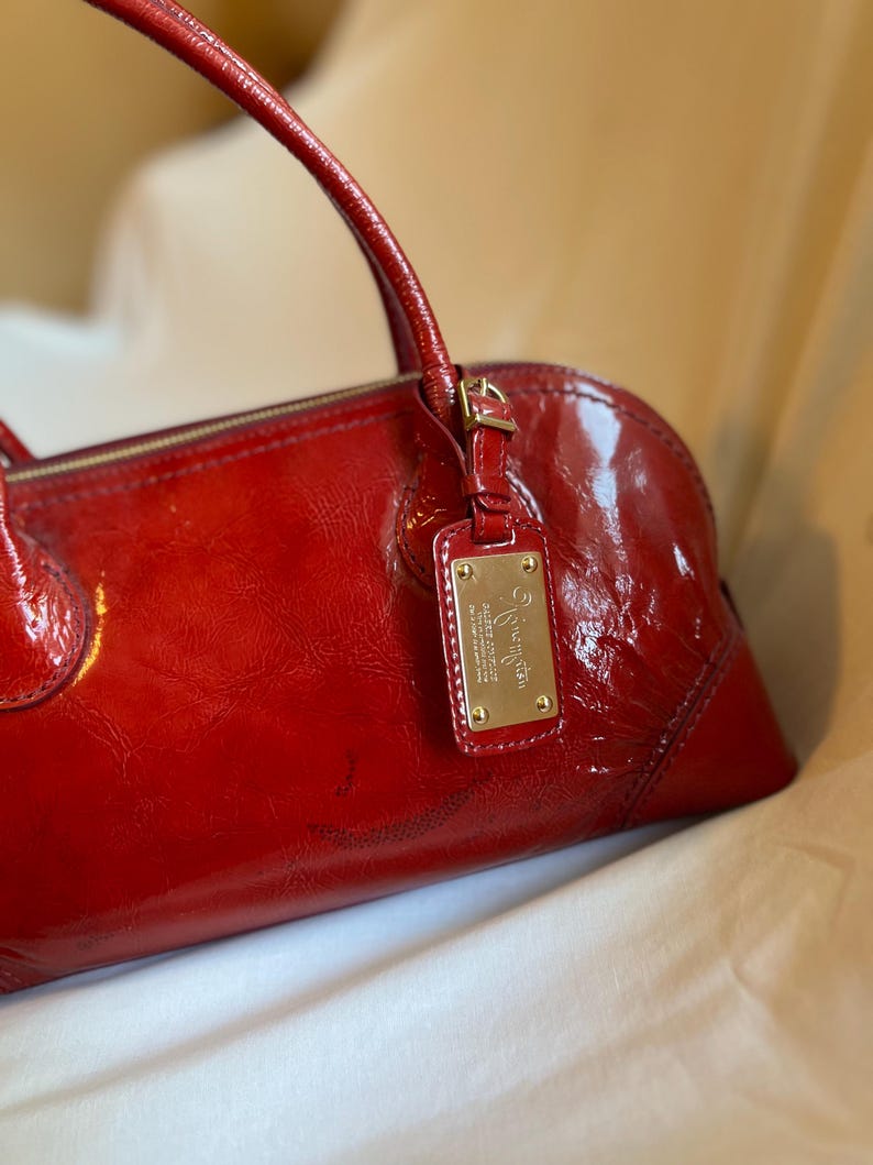 May include: A glossy red handbag with a top handle and a gold-colored metal tag. The bag has a rounded shape and a zipper closure. The tag has text on it.