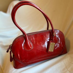 May include: A glossy, red handbag with a rounded shape and two arched handles. The bag features a gold zipper and a small, rectangular tag with gold lettering. The bag's shiny surface reflects light, highlighting its smooth texture.