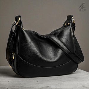 Könnte beinhalten: Eine schwarze Schultertasche aus Leder mit strukturierter Oberfläche. Die Tasche hat ein geschwungenes Design, einen verstellbaren Riemen und goldfarbene Metallbeschläge. Die Tasche liegt auf einer grauen Oberfläche.