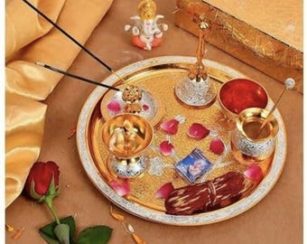 May include: A gold-toned circular metal tray featuring a silver-toned rim, holding a small Ganesha figurine, a bell, a diya lamp, a bowl, a small container, incense sticks, a rectangular card, and a bundle of brown thread, decorated with pink petals.