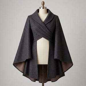 May include: A dark charcoal grey cape coat featuring a wide shawl collar, a wrap-style front with diagonal pleats at the waist, and an open front design. The garment has wide, flowing sleeves and a high-low hemline with a contrasting light brown lining.