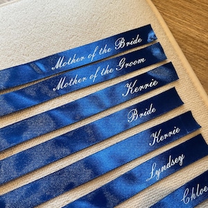 May include: Deep blue satin ribbons with white cursive lettering. The ribbons are inscribed with names and titles including "Mother of the Bride," "Mother of the Groom," and "Bride." The ribbons are laid out on a cream-coloured surface.