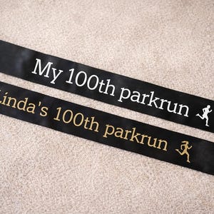 Op de afbeelding: Twee zwarte satijnen linten met witte en gouden tekst. Het bovenste lint leest "My 100th parkrun" met een hardloper-afbeelding. Het onderste lint zegt "Linda's 100th parkrun" met een hardloper-afbeelding. De linten liggen op een beige oppervlak.