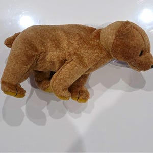 May include: A plush toy shaped like a brown bear in a standing position. The bear has short, soft brown fur, a small rounded tail, and darker brown accents on the bottom of its four paws. It features a small black nose and dark eyes.