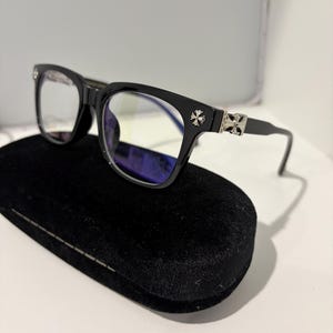 Op de afbeelding: Zwarte bril met rechthoekig montuur en zilveren kruisversieringen op de slapen. De bril ligt op een zwart fluwelen etui. De glazen zijn helder.