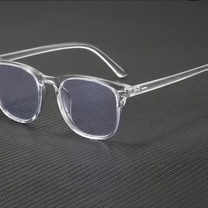 Puede incluir: Gafas con montura transparente y lentes cuadradas. Las gafas tienen una montura y patillas transparentes, y las lentes parecen tener un tinte azul. El diseño es moderno y minimalista.