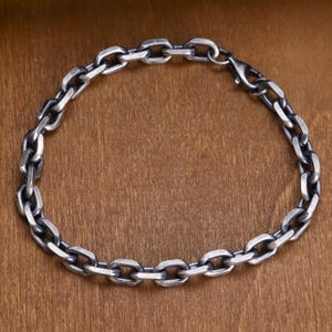 Puede incluir: Un brazalete de cadena plateada con un diseño de eslabones ovalados. El brazalete es de metal y tiene un cierre de broche. El brazalete se muestra sobre una superficie de madera.