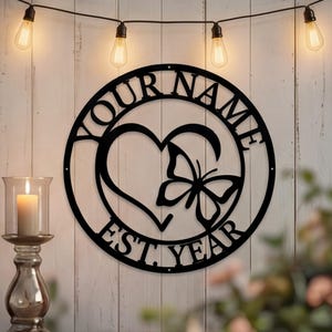 May include: Black metal wall art featuring a heart and butterfly design, with the words "YOUR NAME" and "EST. YEAR" in a circular frame. The art is displayed against a white wooden background, with string lights and a candle holder.
