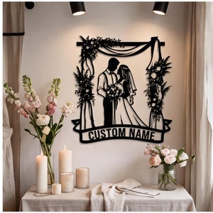 Könnte beinhalten: Ein Wandkunstwerk aus schwarzem Metall, das die Silhouette eines Paares unter einem Blumenbogen zeigt. Das Design enthält ein Banner am unteren Rand mit dem Text CUSTOM NAME. Das Kunstwerk hängt an einer Wand über einem Tisch mit Blumen.
