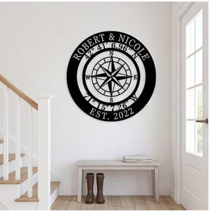 May include: A circular black wall sign featuring a central compass rose design. The text reads ROBERT & NICOLE, the coordinates 42 41 6.96 N and 71 15 7.26 W, and EST. 2022. The piece is constructed with a cutout style, displaying the wall colour through.