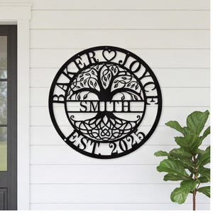 Puede incluir: Un letrero de pared circular de metal negro con un diseño de árbol de la vida. El texto BAKER y JOYCE está arqueado en la parte superior, con un corazón en el centro. El nombre SMITH está centrado sobre el tronco y EST. 2025 en la parte inferior.