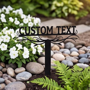 Op de afbeelding: Een zwarte metalen tuinpaal met een horizontaal bord waarop de woorden CUSTOM TEXT in een vetgedrukt schreeflettertype staan. Het bord wordt ondersteund door een verticale paal en is versierd met sierlijke, symmetrische takvormen.