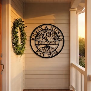 May include: Black metal family name sign with a tree design. The sign is circular with the names "KEVIN & SUSAN" around the top and "BROWN" in the center. The year "EST. 2023" is at the bottom. A green wreath hangs to the left.