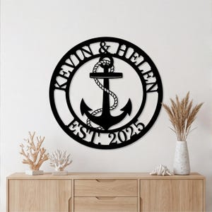 Puede incluir: Un adorno de pared de ancla de metal negro con los nombres "KEVIN & HELEN" y el año "EST. 2025" en un diseño circular. El ancla está detallada con una cuerda y está centrada. La obra de arte está montada en una pared blanca.