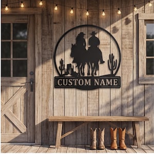 May include: Black metal wall art depicting a cowboy and cowgirl on horseback, set within a circular frame. The design incorporates cacti and a banner reading "CUSTOM NAME". The artwork is displayed on a wooden wall, with a bench and cowboy boots beneath.