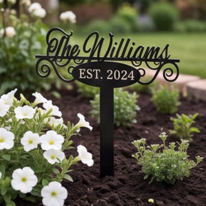May include: A black metal garden stake featuring the text The Williams in a cursive script font above an oval plaque with the text EST. 2024. The sign has decorative scrollwork accents on the sides and a vertical post for ground insertion.