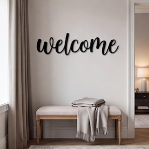 Può includere: Un'insegna da parete nera che riporta la parola welcome in un carattere corsivo. Le lettere sono collegate in un unico pezzo, mostrando una finitura liscia e un aspetto scuro e audace che contrasta con la superficie chiara della parete.