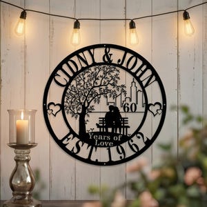 Puede incluir: Arte de pared de metal negro con los nombres "Cidny & John" y el texto "Years of Love EST. 1962". El diseño incluye un árbol, una pareja en un banco, un paisaje urbano y corazones. El arte está iluminado por luces de cadena.