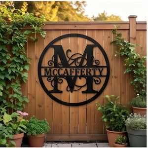 May include: A circular black metal sign featuring a large capital letter M in the center with the name MCCAFFERTY displayed horizontally across the middle. The design includes decorative scrollwork accents within the circular frame.