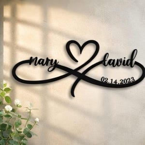 May include: Black metal wall art featuring the names "mary" and "david" intertwined with an infinity symbol and a heart. The date "02.14.2023" is also included. This decorative piece is suitable for weddings or anniversaries.