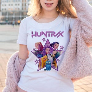 Peut inclure: T-shirt blanc avec le mot "HUNTRX" en violet au-dessus d'un motif en forme d'étoile. Le motif comprend trois personnages féminins stylisés et un chat bleu. Le t-shirt est à manches courtes.