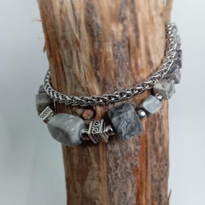 Puede incluir: Una pulsera de varias hebras que presenta una cadena de eslabones tipo trigo en tono plateado y una segunda hebra compuesta por cuentas de piedra gris veteada, intercaladas con pequeñas cuentas espaciadoras y detalles metálicos cuadrados.