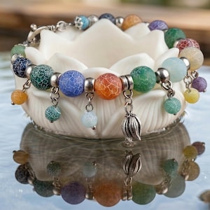 Può includere: Un braccialetto di perline colorate con perline rotonde screpolate nei toni del blu, verde, arancione e viola. Il braccialetto ha dettagli argentati e ciondoli pendenti. È esposto su un supporto bianco a forma di fiore di loto.