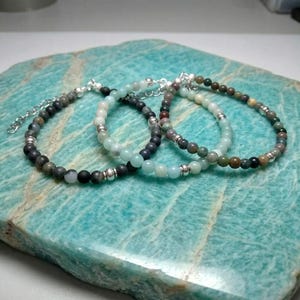 Puede incluir: Tres pulseras de cuentas con una variedad de cuentas en tonos tierra, incluyendo negro, gris y verde, se exhiben sobre una superficie de piedra turquesa. Cada pulsera presenta pequeñas cuentas espaciadoras plateadas y un cierre de cadena para ajustar la talla. Las pulseras son delicadas y elegantes.