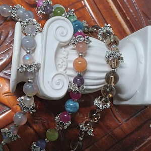Könnte beinhalten: Drei Armbänder mit silberfarbenen Akzenten und verschiedenen Edelsteinperlen, darunter grau, blau, rosa, orange und braun. Die Armbänder werden auf einer weißen dekorativen Säule vor einer braunen Holzoberfläche präsentiert.