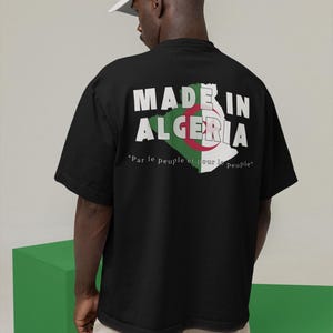 Può includere: Una t-shirt nera a maniche corte con la scritta MADE IN ALGERIA in lettere bianche sopra una grafica della mappa dell'Algeria nei colori verde, bianco e rosso. Sotto la mappa, il testo Par le peuple et pour le peuple è stampato in bianco.
